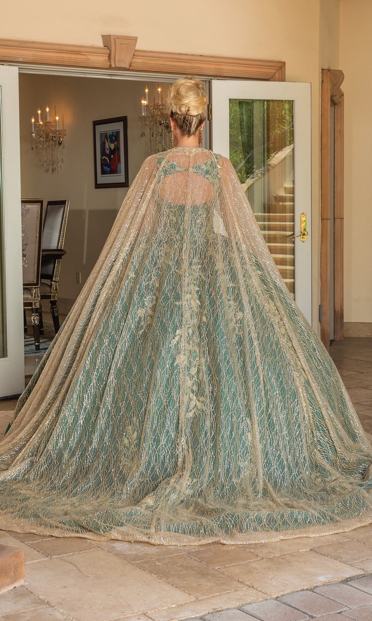 A woman with blonde hair in an updo stands facing away, wearing the Quinceanera Dress 1755 By Dancing Queen, a hunter green gown with gold accents and a sheer embellished cape, inside a building before glass doors.